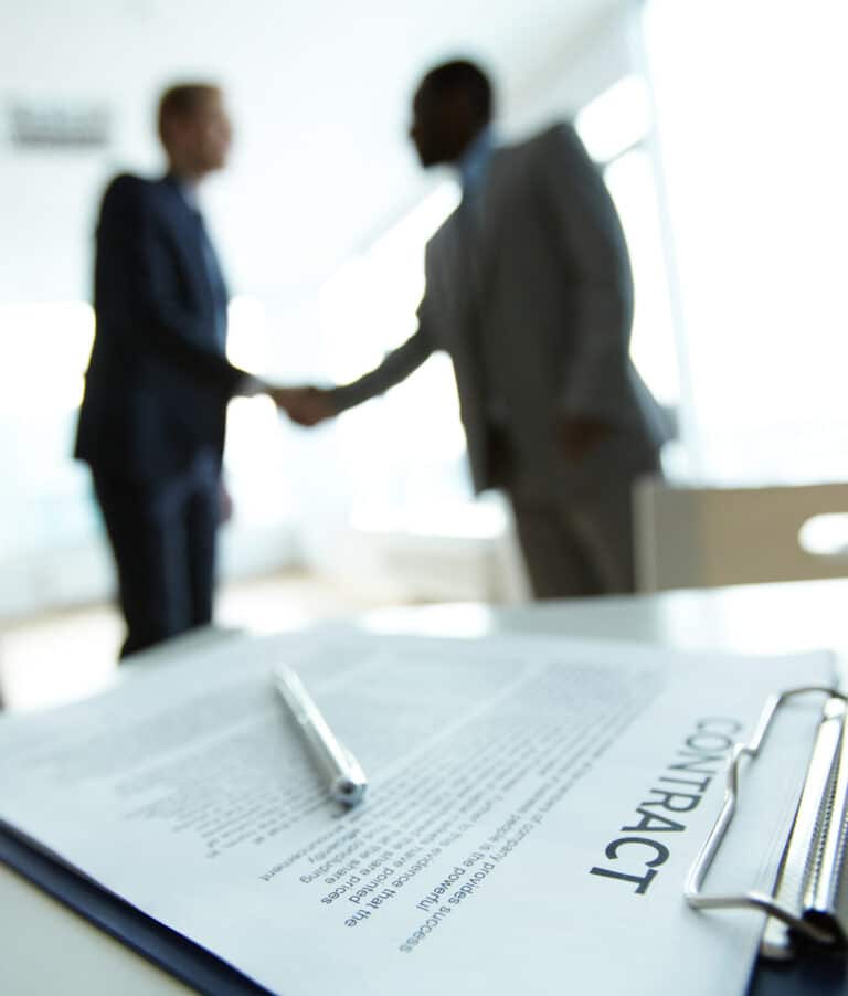 2 men shaking hands in a blurry background. There is a contract with a pen in the forefront.