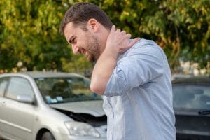 A man holding his neck in pain. There is a wrecked car behind him.
