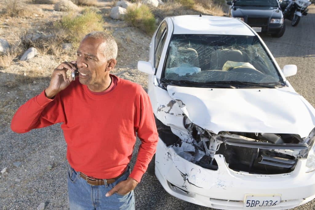 man in red shirt talking on phone with damaged car behind him