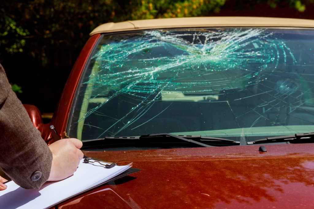 cropped image of insurance adjuster writing on clipboard resting on hood of car with smashed windshield